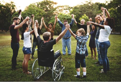 A group holding hands in a circle in a park with one person in a wheelchari