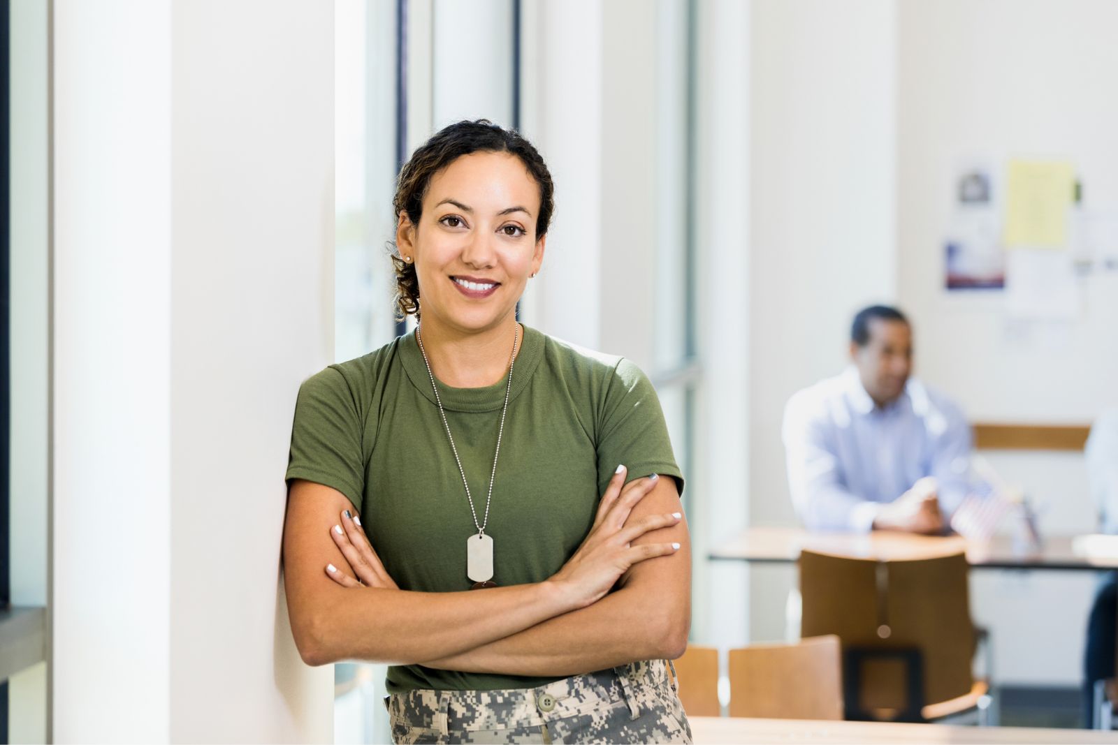 A mid adult female soldier leans against the wall with her arms crossed in an office setting