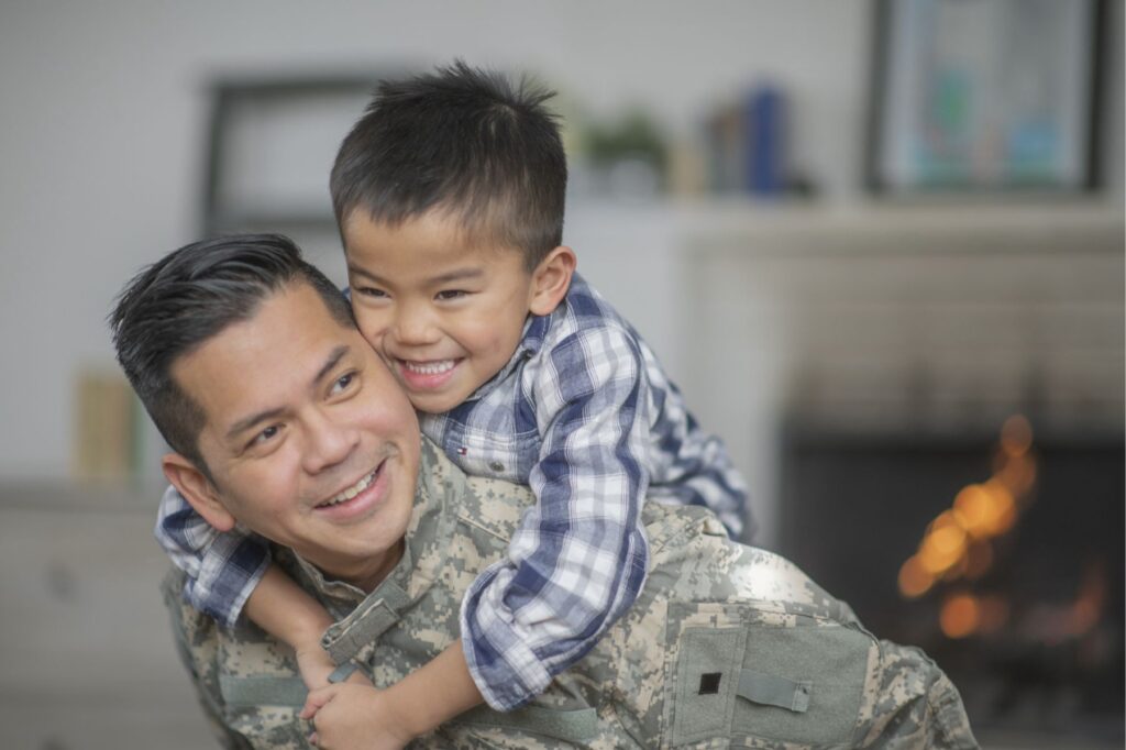 A military dad and his son are hugging in their living room. The son is smiling happily