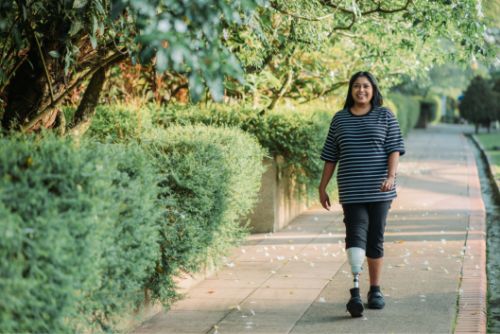 A smiling Latinx woman with a prosthetic leg walks down the sidewalk in her neighborhood surrounded by green trees and bushes.