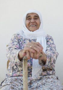 Outdoor portrait of an elderly woman wearing a hijab and holding a cane sitting in front of her house.