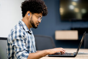 A young black man with glasses and a hearing aid works on a laptop in a conference room. 