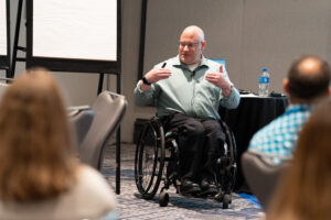 An older man in a wheelchair wearing a light blue button up shirt teaches a class. 
