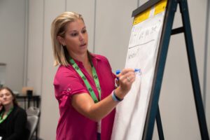 Karma Edwards, NACDD Consultant, is a woman with blonde hair and a pink short sleeve shirt who writes on a large pad in front of a group of people