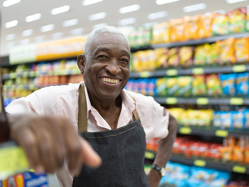 An elderly grocery store worker rests his arm smiling with an aisle of food behind him.