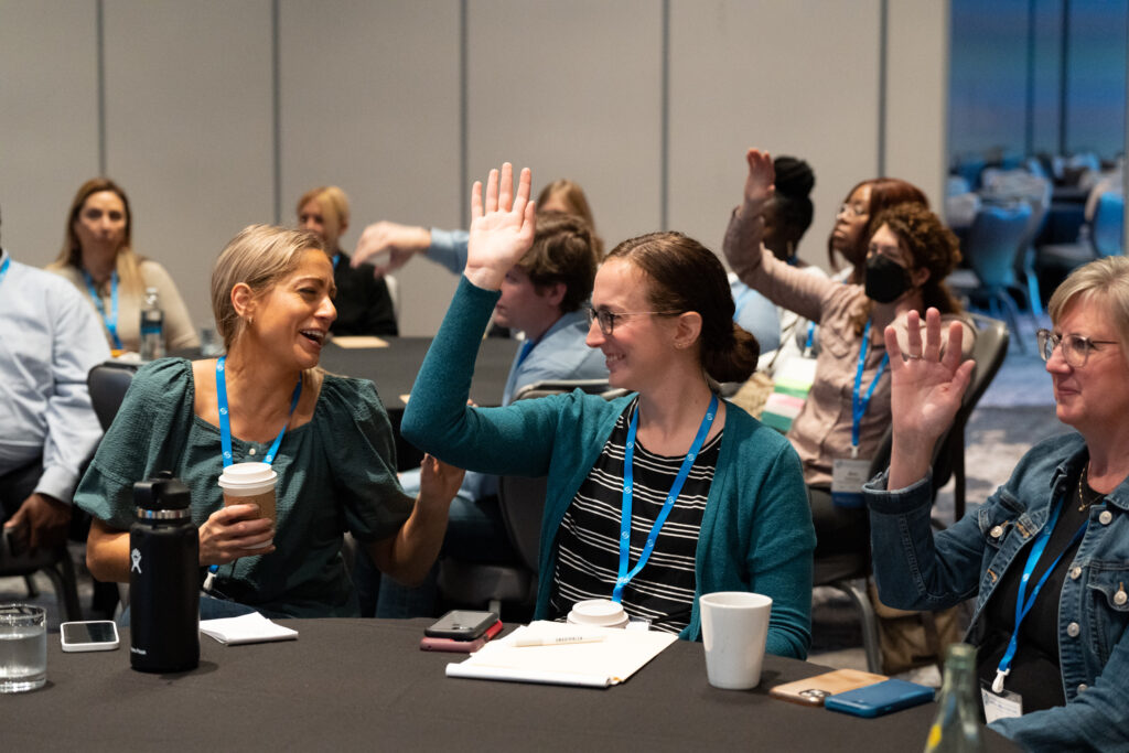 Three women sitting at a table in a large conference room. Two of the three women are raising their hands. The woman on the left is laughing with the woman in the middle. There are multiple other people in the background.