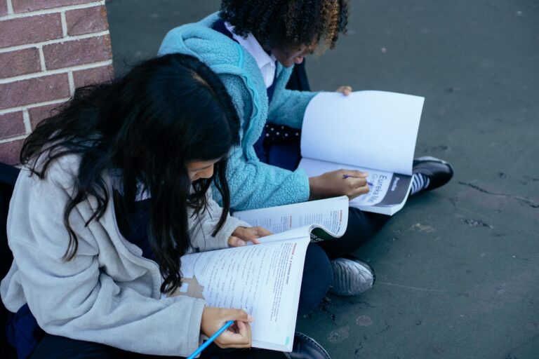 Two students sit on the ground outside against a brick wall, focused on reading and writing in their workbooks.