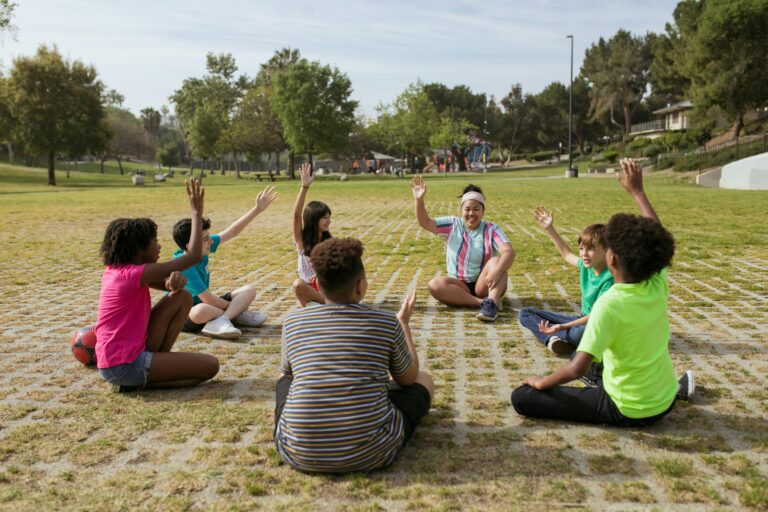 A group of children and a young adult sit in a circle on a grassy field, raising their hands and smiling during a team-building activity.