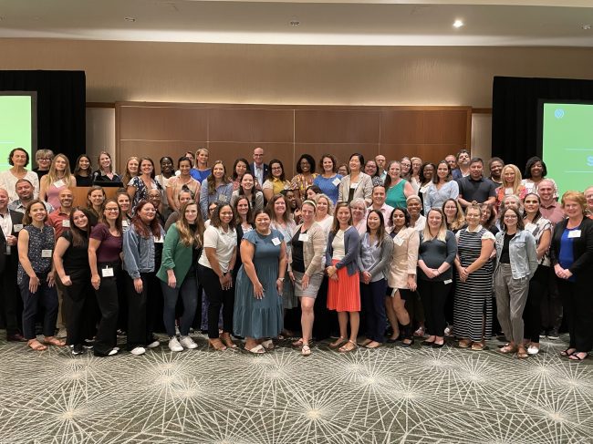 A group photo of panelists and attendees of the Summit to Advance Healthy Communities, Reflecting, Sharing, and Planning for the Future. There is a large group of people arranged for a group photo with multiple rows of people. They are in a large event space with two large display screens on either side of the group