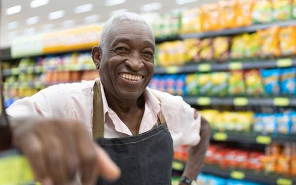 An elderly grocery store worker rests his arm smiling with an aisle of food behind him.