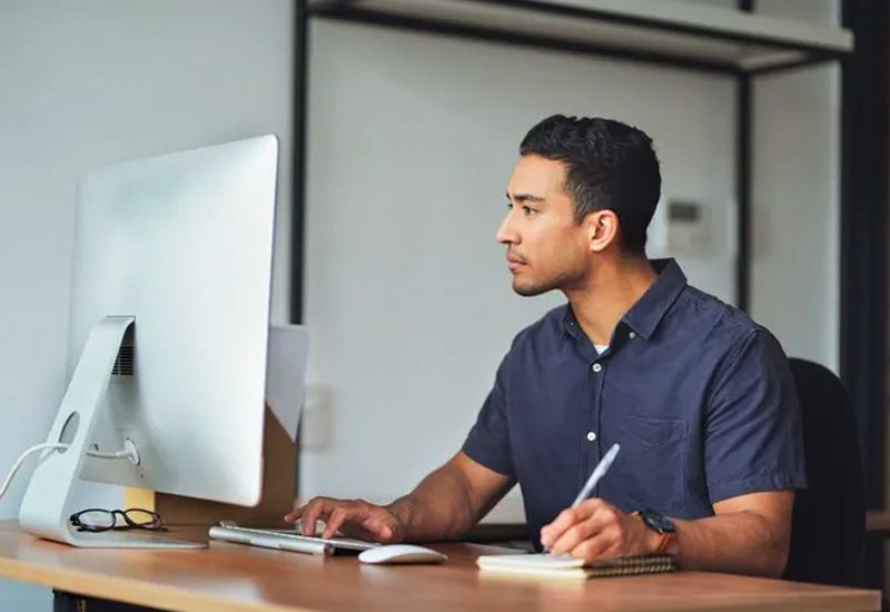 A man writing on a notepad while looking at computer screen.