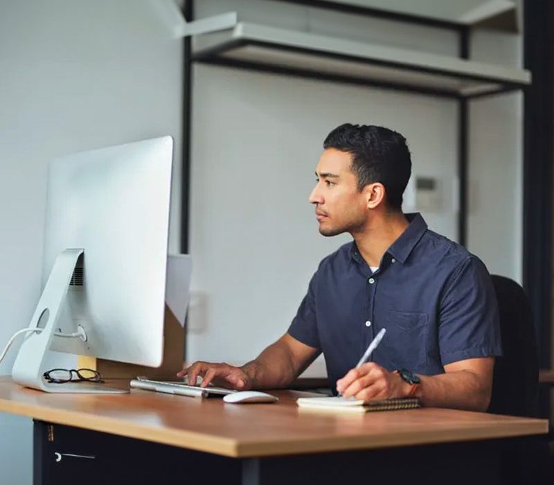 A man writing on a notepad while looking at computer screen.
